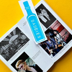 Puede incluir: Un libro abierto con un marcapáginas azul que dice "DADDIES". El libro tiene varias fotos de un hombre en diferentes entornos. El texto en la página dice "May 1976: David fans the flames over his rumoured obsession with Hitler with an apparent Nazi salute at Victoria station, London." y "Striking a pose off-set during filming of The Man Who Fell to Earth, New Mexico, 1975."