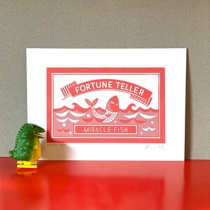 May include: A red and white print of a fish with the text "Fortune Teller" and "Miracle Fish" in a red frame. The print is signed "yell" in the bottom right corner.
