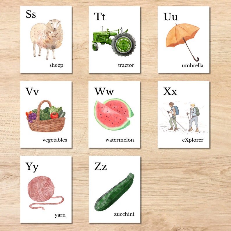 May include: Set of watercolor alphabet flashcards. Each card features a letter and a corresponding image: a sheep, tractor, umbrella, vegetables, watermelon, explorers, yarn, and zucchini. The cards are on a wooden surface.