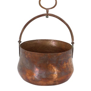 May include: A hammered copper cauldron with a rounded base and a curved handle. The handle is attached to the rim with rivets and has a circular loop at the top. The copper has a rich, warm color and a textured surface.