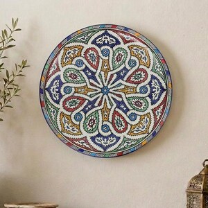 May include: A decorative, round plate with a colourful, intricate pattern of blue, red, yellow, and green. The plate is mounted on a beige wall above a wooden table. A woven basket with a plant and a decorative lantern are also on the table.