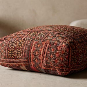 Könnte beinhalten: Ein quadratisches Bodenkissen mit einem komplizierten, geometrischen Muster in Rot-, Braun- und Schwarztönen. Das Kissen hat ein strukturiertes Aussehen und ist wahrscheinlich aus Stoff. Das Design deutet auf eine Boho- oder global inspirierte Ästhetik hin.