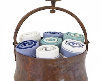 May include: A rustic copper-toned metal basket with a handle, filled with rolled-up towels. The towels are in various shades of blue, white, and teal, with striped and solid patterns. The basket has a hammered texture and a curved handle.