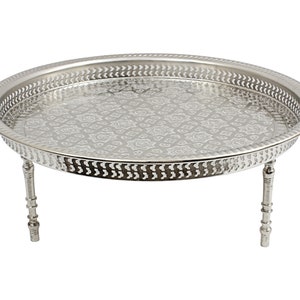 May include: A silver-coloured, round metal tray with a decorative floral pattern on the surface. The tray has a pierced border and is supported by three short, decorative legs. The tray is designed for serving or display.