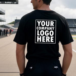 May include: Black t-shirt with the text "YOUR COMPANY LOGO HERE" in a white box. The person is standing on a racetrack. The shirt is short-sleeved.