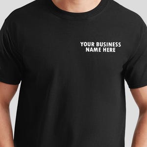 May include: Black t-shirt with the text "YOUR BUSINESS NAME HERE" printed in white on the upper chest. The shirt has a crew neck and short sleeves. The person is wearing the shirt.