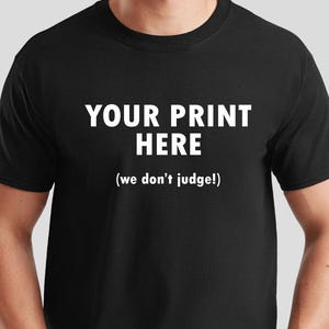 May include: Black crew neck t-shirt with the text "YOUR PRINT HERE" in white, with the phrase "(we don't judge!)" below. The shirt is a solid black color.
