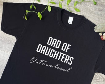 May include: A black short-sleeved t-shirt featuring the white text DAD OF DAUGHTERS in a bold sans-serif font, with the word Outnumbered written below in a white cursive script. The shirt has a standard crew neckline and a white inner label.