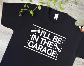 May include: A black short-sleeved t-shirt featuring white text that reads "I'LL BE IN THE GARAGE" centered on the chest. The design includes graphic icons of a hammer and a wrench, along with small nail symbols, all printed in white ink.