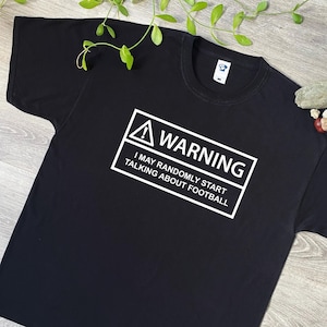 May include: Black t-shirt with a white warning sign that says "WARNING I MAY RANDOMLY START TALKING ABOUT FOOTBALL"