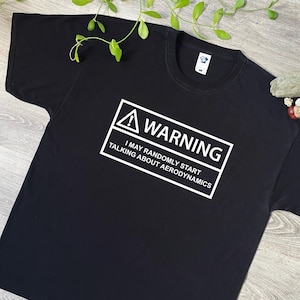 Puede incluir: Camiseta negra con un cartel de advertencia blanco que dice "ADVERTENCIA PUEDO EMPEZAR A HABLAR DE AERODINÁMICA ALEATORIAMENTE"