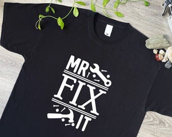 May include: A black short-sleeved t-shirt featuring white screen-printed text that reads MR. FIX IT. The design includes graphic illustrations of a wrench, a hammer, and several nails arranged around the bold, capitalized lettering.