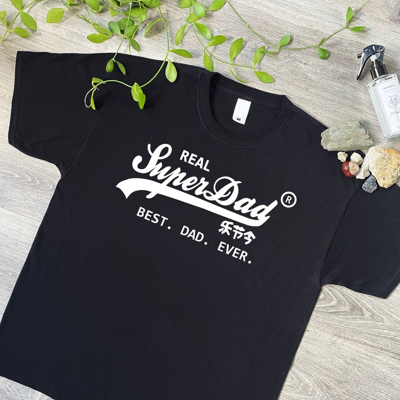 May include: A black short-sleeved t-shirt featuring white screen-printed text that reads REAL SuperDad with a registered trademark symbol, followed by the text BEST. DAD. EVER. and Chinese characters below the word SuperDad.