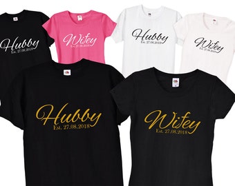 Może przedstawiać: Pięć koszulek z napisami "Hubby" i "Wifey" w ozdobnym, pochylonym kroju pisma. Koszulki są czarne, białe, różowe i szare. Tekst na koszulkach brzmi "Hubby Est. 27.08.2018" i "Wifey Est. 27.08.2018".
