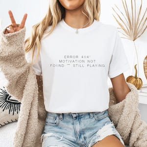 May include: A white short-sleeved t-shirt featuring the black printed text ERROR 404: MOTIVATION NOT FOUND — STILL PLAYING. The shirt has a crew neckline and is worn with a beige textured cardigan and light blue denim shorts with frayed hems.