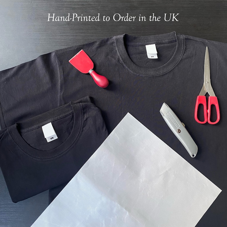 May include: Two black t-shirts, a red scraper, a silver utility knife, a pair of red scissors, and a sheet of white paper on a black surface.  Hand-Printed to Order in the UK.