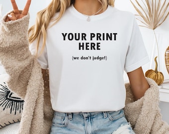 May include: White t-shirt with the text "YOUR PRINT HERE" in bold black letters, with the phrase "(we don't judge!)" underneath. The model is wearing blue denim shorts and a beige jacket.