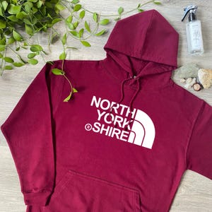 May include: A maroon long-sleeve hooded sweatshirt featuring a white graphic print on the chest that reads NORTH YORK SHIRE with a registered trademark symbol and a stylized mountain-like arc design. The hoodie includes a front kangaroo pocket.