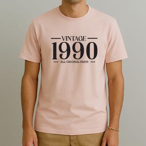 Puede incluir: Camiseta rosa claro con el texto negro "VINTAGE 1990 ALL ORIGINAL PARTS". La camiseta es de manga corta y tiene cuello redondo. La persona lleva una pulsera dorada.