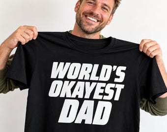 May include: A black short-sleeved t-shirt featuring the white text "WORLD'S OKAYEST DAD" printed in a bold, sans-serif font across the chest. The garment has a classic crew neckline and a standard fit, showcasing clear, legible lettering on the front.