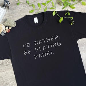 Peut inclure: Un t-shirt noir à manches courtes avec le texte blanc "I'D RATHER BE PLAYING PADEL" imprimé sur le devant. Le vêtement présente une encolure ronde et une étiquette de marque blanche visible au niveau du col intérieur.