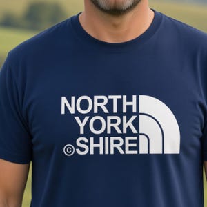 Op de afbeelding: Een marineblauw t-shirt met korte mouwen en een witte grafische print op de borst. Het ontwerp bestaat uit de gestapelde tekst NORTH YORK SHIRE met een copyright-symbool en een gestileerde kwartcirkelvorm bestaande uit drie verticale lijnen.