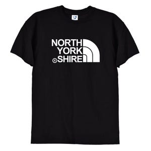 May include: A black short-sleeved t-shirt featuring a white graphic print on the chest. The design consists of the text NORTH YORK SHIRE stacked vertically with a registered trademark symbol and a stylized white quarter-circle shape to the right.