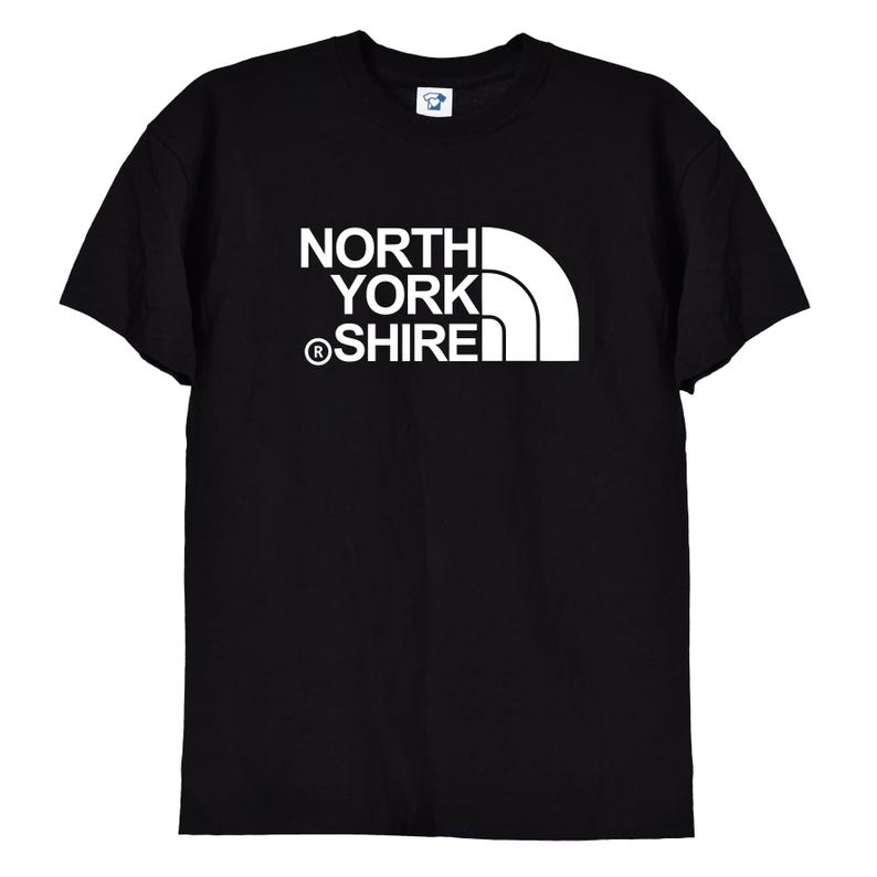 May include: A black short-sleeved t-shirt featuring a white graphic print on the chest. The design consists of the text NORTH YORK SHIRE stacked vertically with a registered trademark symbol and a stylized white quarter-circle shape to the right.