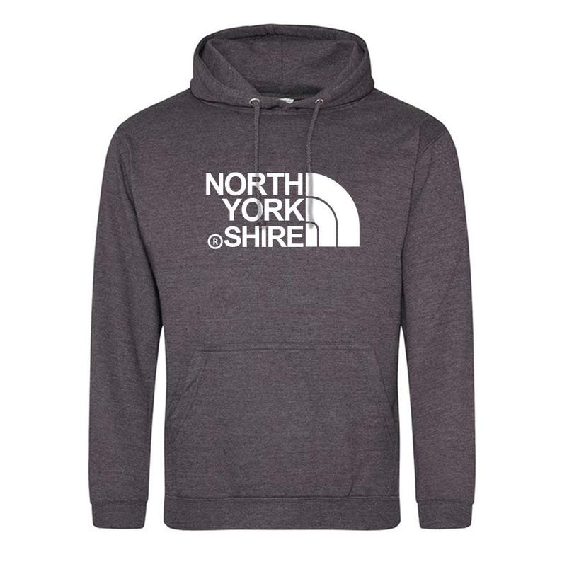May include: A dark charcoal heather grey hooded sweatshirt featuring a white graphic print on the chest. The text reads NORTH YORK SHIRE with a registered trademark symbol and a stylized white quarter-circle graphic design element.