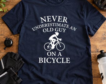 May include: A navy blue short-sleeve t-shirt featuring white text that reads NEVER UNDERESTIMATE AN OLD GUY ON A BICYCLE, accompanied by a white graphic illustration of a person riding a bicycle in the center of the chest.