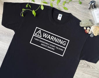 May include: A black short-sleeved t-shirt featuring a white rectangular graphic with a warning symbol and the text: WARNING I MAY RANDOMLY START TALKING ABOUT BAKING. The shirt has a crew neckline and a white interior label at the collar.