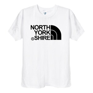 May include: A white short-sleeved t-shirt featuring a black graphic print on the chest. The design consists of the text NORTH YORK SHIRE with a registered trademark symbol and a stylised black quarter-circle shape positioned to the right side.
