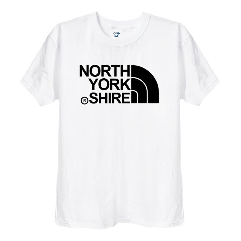 May include: A white short-sleeved t-shirt featuring a black graphic print on the chest. The design consists of the text NORTH YORK SHIRE with a registered trademark symbol and a stylised black quarter-circle shape positioned to the right side.