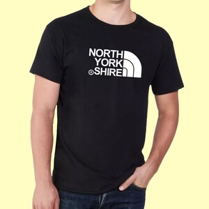 May include: A black short-sleeved t-shirt featuring a white graphic print on the chest. The design consists of the text NORTH YORK SHIRE stacked vertically with a registered trademark symbol and a stylized white quarter-circle shape to the right.