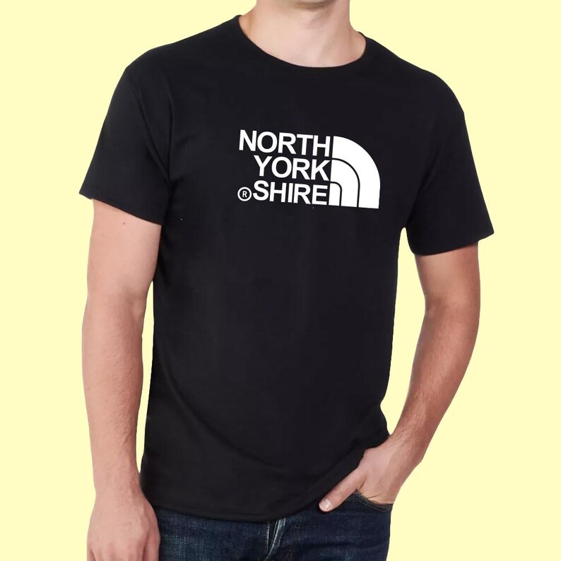 May include: A black short-sleeved t-shirt featuring a white graphic print on the chest. The design consists of the text NORTH YORK SHIRE stacked vertically with a registered trademark symbol and a stylized white quarter-circle shape to the right.