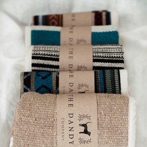May include: Five rolled blankets with patterned woven designs in various colours, including teal, black, and brown. Each blanket is wrapped with a brown paper band printed with "THE DANDY DOG COMPANY" and a dog silhouette.
