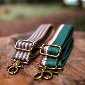 Puede incluir: Dos correas de bolso a rayas con herrajes color latón. Una correa es beige con rayas azul marino, la otra es verde con rayas blancas y burdeos. Ambas tienen cierres giratorios.