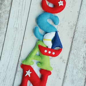 May include: A felt letter banner spelling out the name "SAND", with each letter decorated with a different vehicle. The "S" has a white airplane, the "A" has a blue sailboat, the "N" has a red car, and the "D" has a blue car.