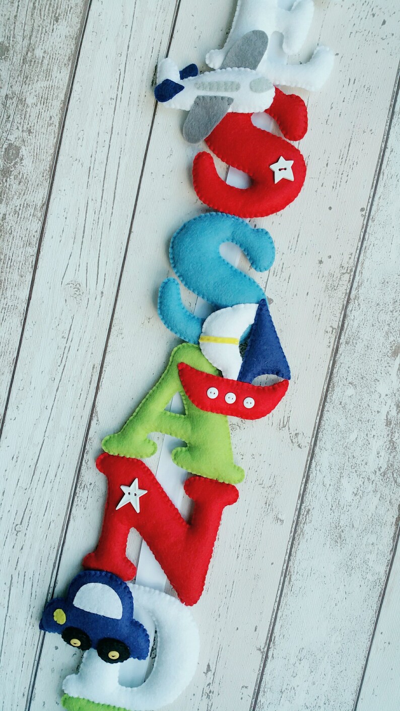 May include: A felt letter banner spelling out the name "SAND", with each letter decorated with a different vehicle. The "S" has a white airplane, the "A" has a blue sailboat, the "N" has a red car, and the "D" has a blue car.