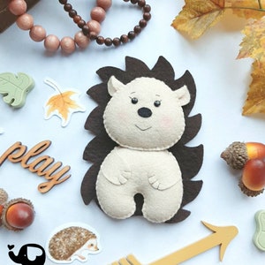 May include: A handmade felt hedgehog with a cream-colored body and brown spines. The hedgehog is surrounded by autumn-themed decorations, including acorns, leaves, and a wooden arrow. The image also includes the text "play" and the logo for "The Little Elephant".