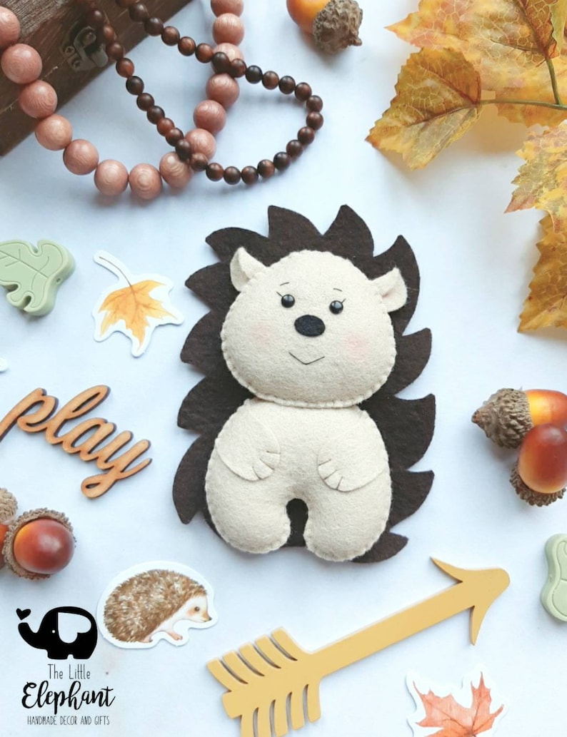 May include: A handmade felt hedgehog with a cream-colored body and brown spines. The hedgehog is surrounded by autumn-themed decorations, including acorns, leaves, and a wooden arrow. The image also includes the text "play" and the logo for "The Little Elephant".