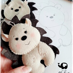 May include: Handmade felt hedgehog ornaments in cream and brown. The hedgehogs have black eyes, pink cheeks, and dark brown spines. One is complete, and another is in progress with a pin. A pencil sketch of a hedgehog is visible in the background.