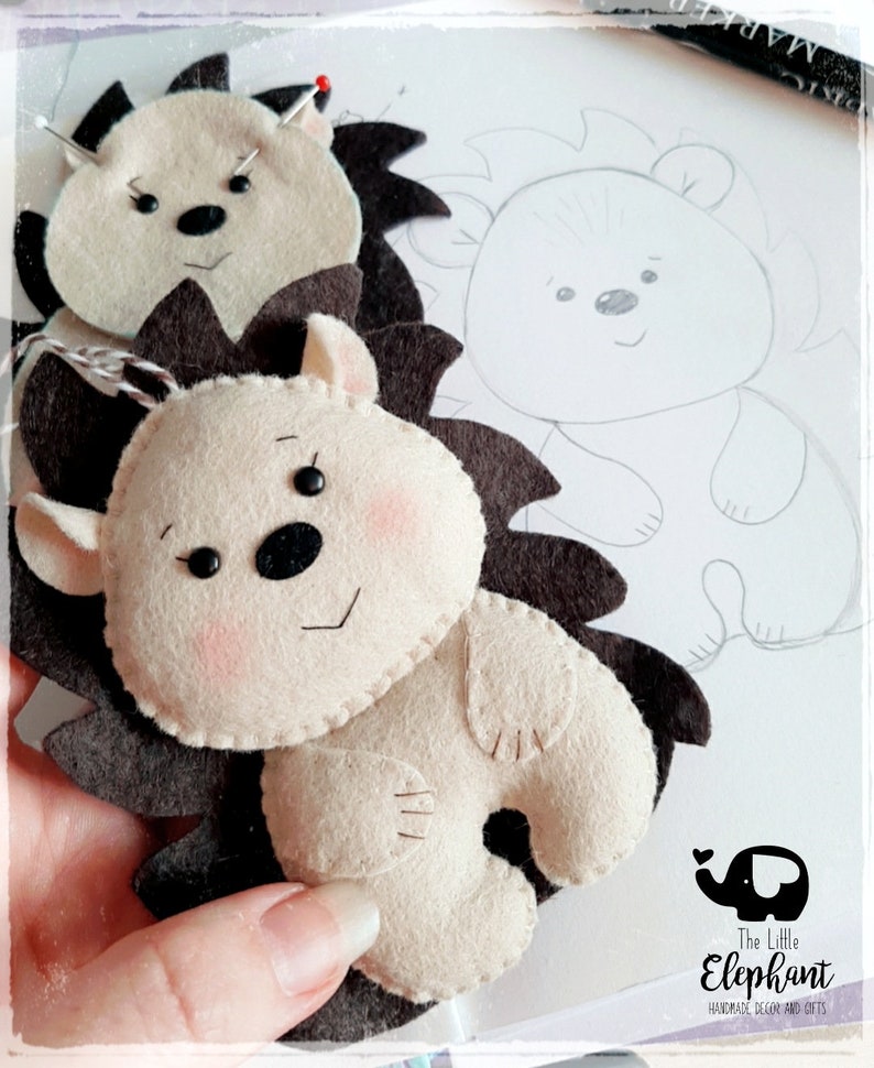 May include: Handmade felt hedgehog ornaments in cream and brown. The hedgehogs have black eyes, pink cheeks, and dark brown spines. One is complete, and another is in progress with a pin. A pencil sketch of a hedgehog is visible in the background.