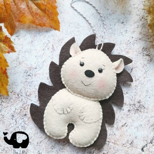 May include: A handmade felt hedgehog ornament with a cream-colored body, brown spines, and a string for hanging. The hedgehog has a smiling face with pink cheeks and a black nose. The Little Elephant logo is in the bottom left corner.