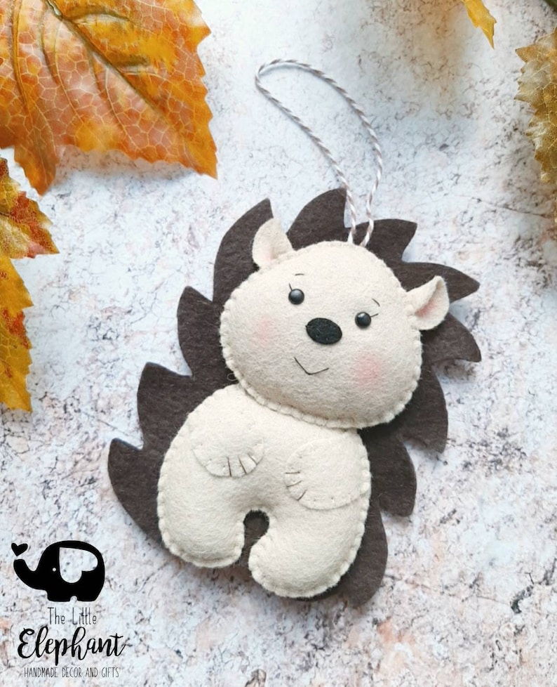 May include: A handmade felt hedgehog ornament with a cream-colored body, brown spines, and a string for hanging. The hedgehog has a smiling face with pink cheeks and a black nose. The Little Elephant logo is in the bottom left corner.