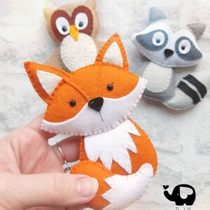 May include: Felt animal ornaments, including a fox, owl, and raccoon. The fox is orange with white accents, the owl is brown with white eyes, and the raccoon is gray with black stripes. The ornaments are handmade and feature embroidered details.