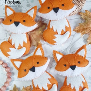 May include: Four orange felt fox ornaments with white accents. The foxes have black eyes and noses. The foxes are hanging from a string.
