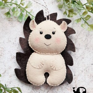 May include: A handmade felt hedgehog ornament with a cream-colored body, brown quills, and pink cheeks. The ornament has a string for hanging and is set against a light background with greenery. The Little Elephant logo is visible.