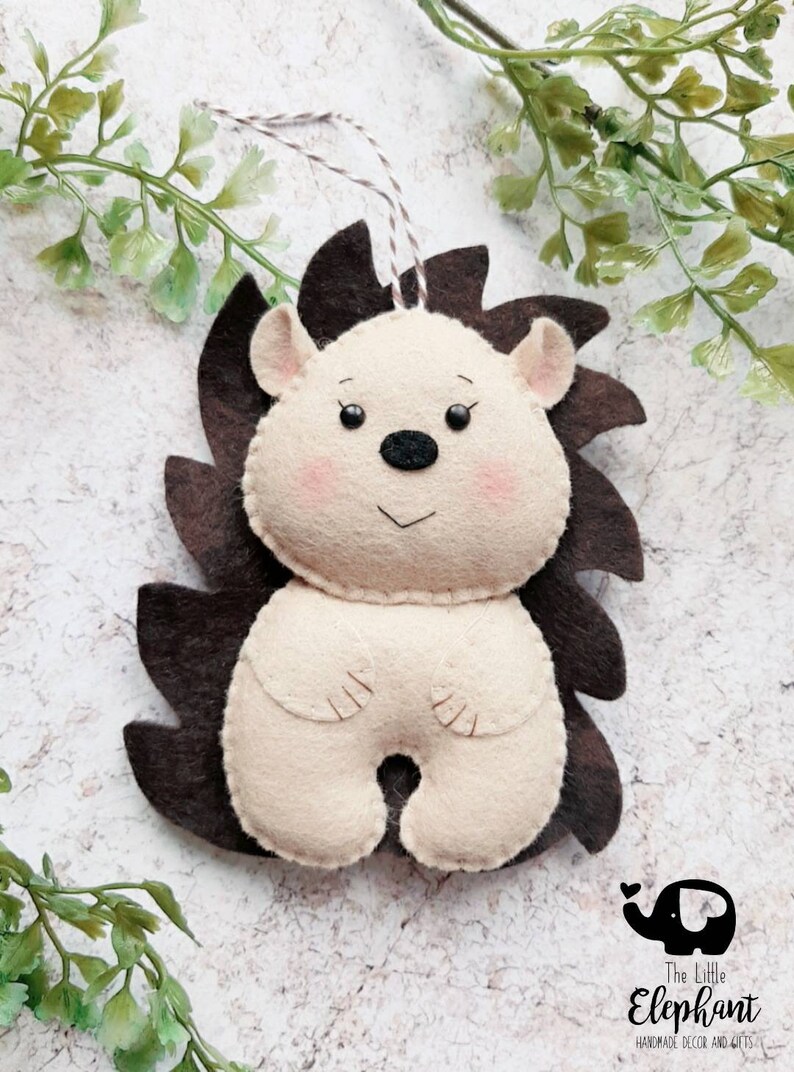 May include: A handmade felt hedgehog ornament with a cream-colored body, brown quills, and pink cheeks. The ornament has a string for hanging and is set against a light background with greenery. The Little Elephant logo is visible.