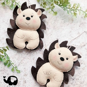 May include: Two felt hedgehog ornaments with brown spines and cream-colored bodies. Each has a smiling face with pink cheeks, black eyes, and a black nose. They hang from white string. The Little Elephant logo is in the lower left.